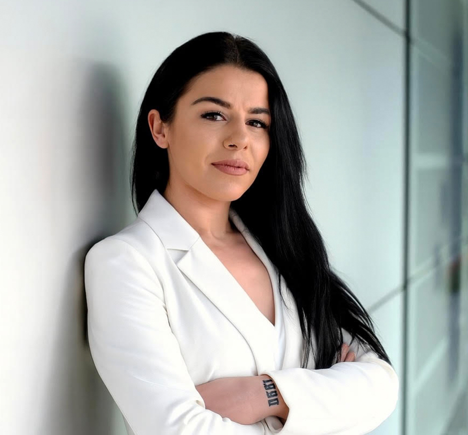 Kübra, eine selbstbewusste Mitarbeiterin in einem weißen Blazer, lächelt leicht, während sie sich an eine Wand lehnt. Ihr Ausdruck spiegelt die Wertschätzung für ihr unterstützendes Arbeitsumfeld wider.